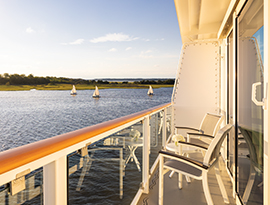 The private balcony on the American Pioneer, furnished with chairs and a small table, overlooking a sun-drenched waterway filled with several small sailboats near a grassy shoreline.