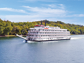 White river cruise ship sails on calm river with green forested shore in background, clear sky above.