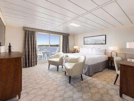 A spacious cruise ship suite featuring a large bed, two armchairs, and sliding glass doors that open to a private balcony overlooking Alaskan waters.