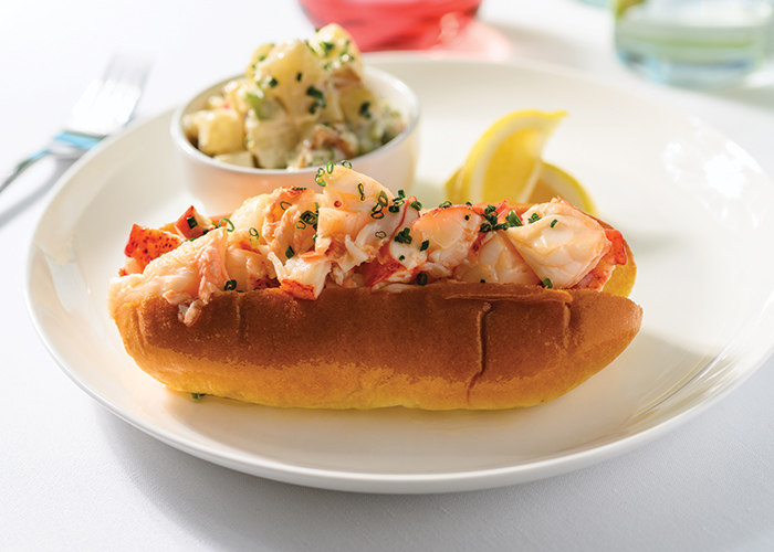 A dish containing a fresh lobster roll, lemon slices, and a small bowl of potato salad, served fresh on American Cruise Lines.