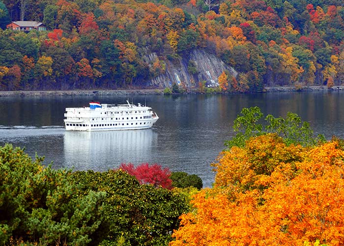 An American Cruise Lines Coastal Cat small cruise ship sailing along the Hudson River surrounded by cliffs with vibrant fall foliage trees on each side of the river.