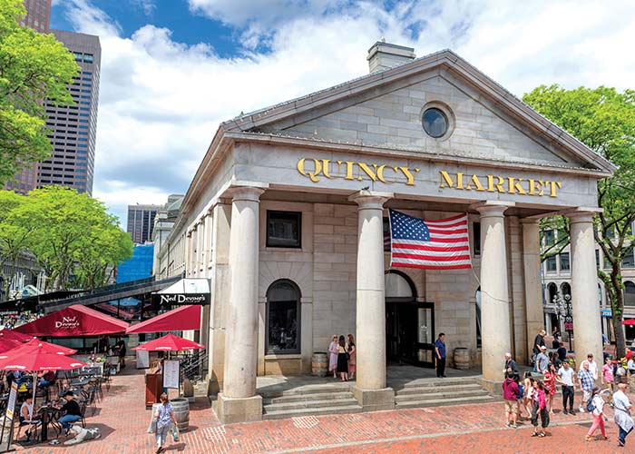 Quincy Market, a light brick building in Boston with four pillars in the front and an American flag between the two in the middle, and several chairs and tables with red umbrellas to the left of the building. A gold sign with Quincy Market written on it is above the entryway, and there are several people walking outside the building.