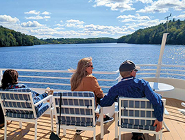 Three cruise guests sitting on blue and white striped chairs on the sun deck of American Eagle, an American Cruise Lines Coastal Cat small cruise ship, overlooking an intimate waterway with dense forest-land on each side of the water.