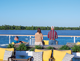 Three people relax on a river cruise ship deck with yellow chairs, overlooking calm water and green shoreline.