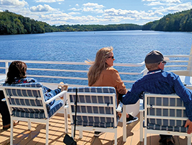 Three people relax on a cruise ship deck overlooking a calm river with forested banks under a blue sky.