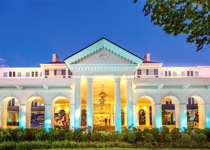 The Guest House at Graceland, a bright white building with elegant columns that lead to a roof-covered drop-off area in Memphis. There are blue lights shining from the ground up on the white columns, covered by small, trimmed bushed lining the front of the building.