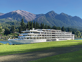 A modern riverboat positioned close to a lush green riverbank, highlighting a scenic destination stop beneath the mountain peaks of the Columbia River Gorge.