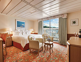 The Veranda Suite on American Constellation small cruise ship, with floor-to-ceiling sliding glass doors that lead to a private balcony with a table and chairs overlooking the Inside Passage. There is a bed with a bedside table and a lamp on top of it on each side, and two chairs by the foot of the bed, all over a red-patterned rug. The bed is facing a wooden dresser with a TV on the wall above it