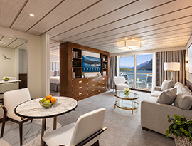 A spacious ship stateroom on American Encore featuring a small dining table, a living area with a sofa and television, and sliding glass doors that open to a private balcony overlooking a scenic river.