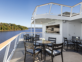 The outdoor Back Porch Café on American Constellation featuring tables and chairs on the deck, offering scenic views of a tree-lined river.