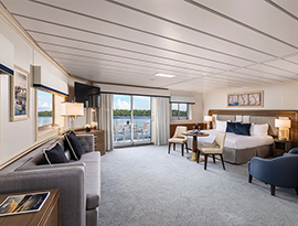 The spacious Veranda Suite on American Heritage, with floor-to-ceiling sliding glass doors that lead to a private balcony with a table and chairs overlooking the Mississippi River. There is a bed on the right side of the room and an end table with a lamp on each side of the bed. At the foot of the bed, there are two chairs facing a TV on the wall above a wooden dresser in the corner.