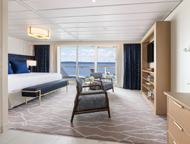 A spacious luxury stateroom on a modern cruise ship, featuring a large bed with deep blue accents, a private sitting area with two upholstered armchairs, and floor-to-ceiling glass doors that open to a balcony overlooking the open water.
