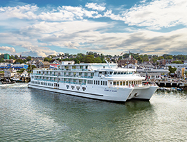 The modern multi-deck coastal cruise ship American Eagle navigating calm harbor waters alongside a scenic waterfront town under a partly cloudy sky.