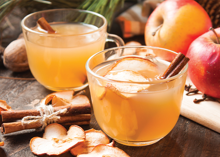 On a grey wooden surface, there are two glasses of light orange colored cider with thin apple slices and a cinnamon stick in each, with a few thin apple slices scattered around the glasses, and two red apples in the background.