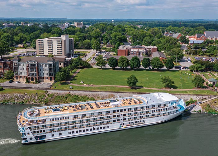 The American Riverboat American Symphony docked along the Tennessee River with its retractable bow gangway letting passengers off the ship in Decatur, with an aerial view of the sun deck on the top of the ship. On land, there is a large empty grass area surrounded by a few buildings and trees.