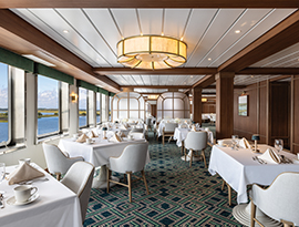 The glass-enclosed restaurant on American Cruise Lines' American Patriot, featuring tables with white linens and panoramic window views of the spectacular scenery.