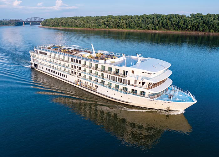 The American Serenade, an American Cruise Lines Riverboat gliding along the Mississippi River, with the tree-filled shore and a bridge in the background.