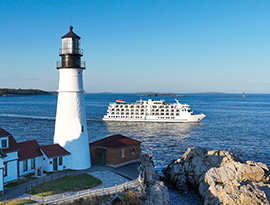 A scenic view of a classic white lighthouse and a red-roofed keeper's house on a rugged, rocky shoreline, with a modern multi-deck cruise ship sailing on the blue ocean under a clear sky.