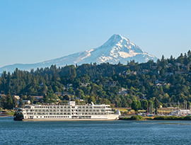 The American Jazz cruises along a waterway near a lush, tree-lined town, set against the breathtaking, snow-capped backdrop of a majestic mountain.