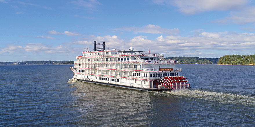 White paddlewheel river cruise ship sailing on a wide river, green shoreline and blue sky in background.