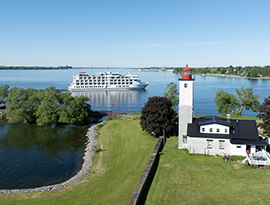 An aerial view of the cruise ship American Patriot passing a historic white lighthouse with a red top on a grassy shoreline.