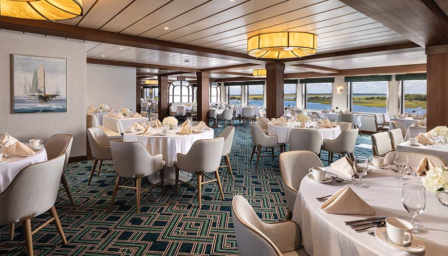 Elegant dining room on board an American Cruise Lines ship with round tables, white linens, large windows showing river and green landscape, modern decor, and nautical decor.