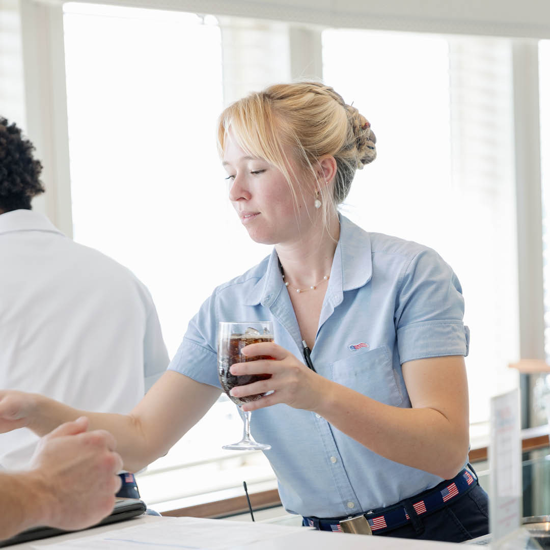 A server in a blue American Cruise Lines uniform hands a drink to a guest on board.