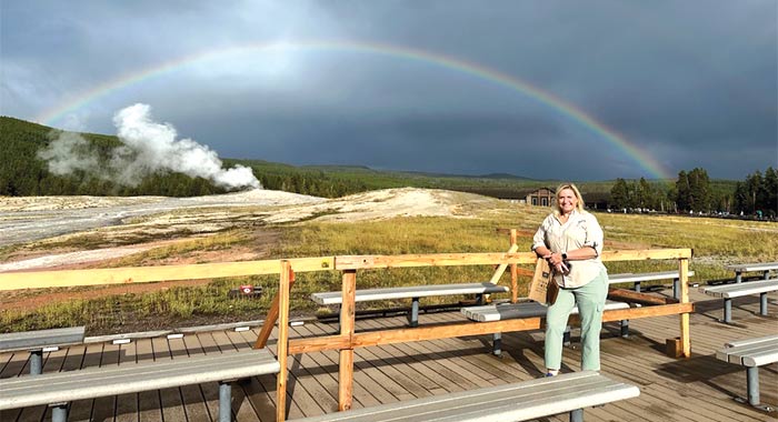 An American Cruise Lines guest stands on wooden deck with benches at Yellowstone National Park, with a rainbow and geyser in background.