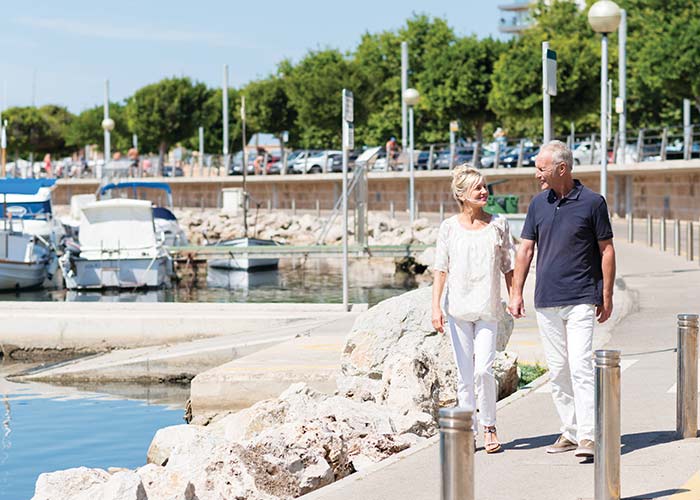 Elderly couple walking along the Newport marina during a cruise stop, with boats and trees in the background
