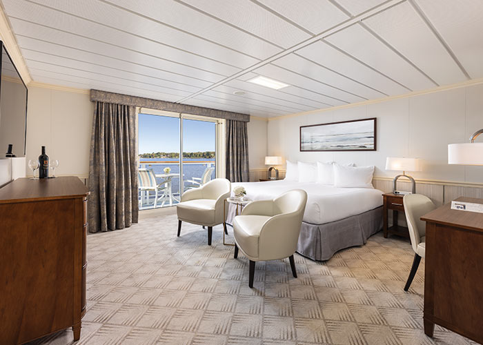 Luxurious American Cruise Lines cabin with a large bed, seating area, and balcony overlooking a river. Elegant decor includes a landscape painting, wine bottle, and scenic view.