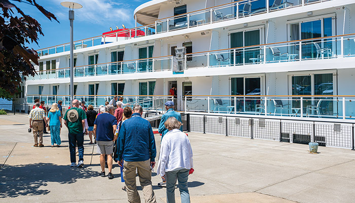 Guests boarding a small American Cruise Lines river ship with private stateroom balconies, docked at a sunny port.