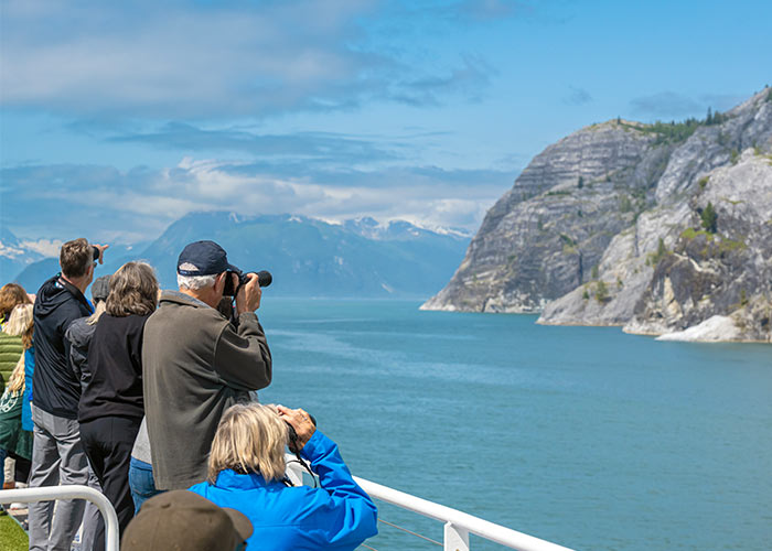 People on a Constellation Class cruise ship deck photographing scenic mountains and water.