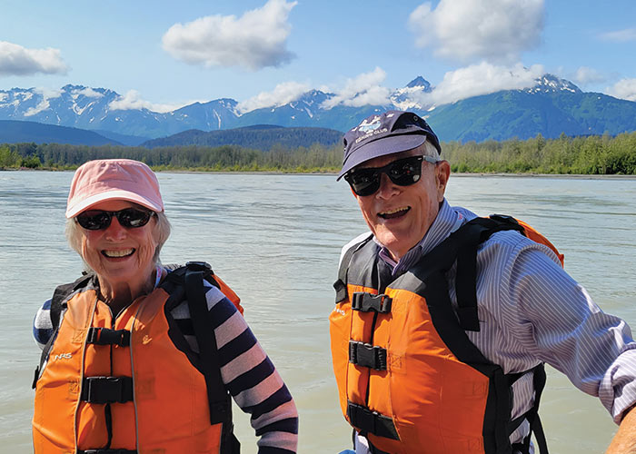 Couple in life vests smiling on a river cruise with American Cruise Lines, snowy mountains and forest in the background.