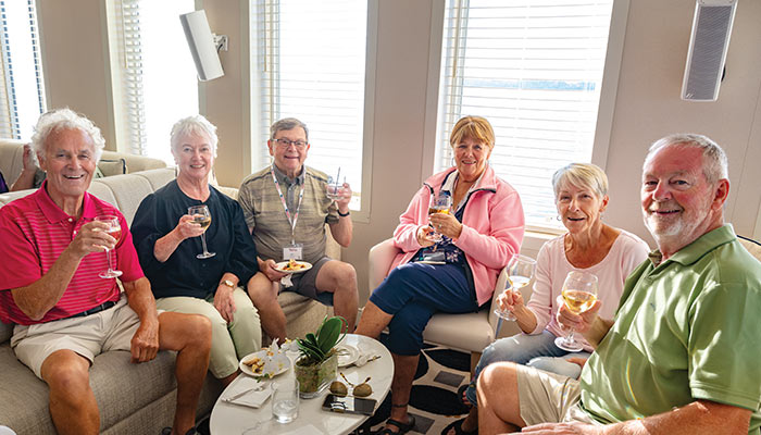 A group of six older adults enjoying wine and snacks in a cozy lounge on an American Cruise Lines ship, with large windows showing a river view.