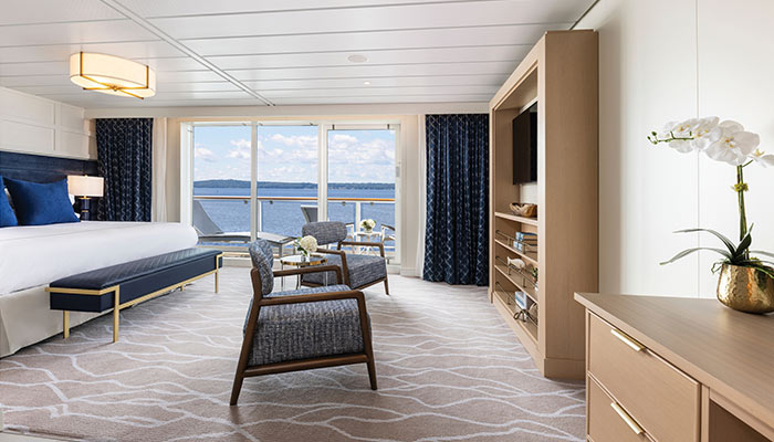 Luxurious American Cruise Lines ship stateroom with a large bed, seating area, TV, and private balcony overlooking a scenic river.