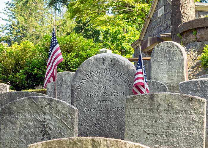 Gravestones with American flags, surrounded by trees, in the Sleepy Hollow Cemetery.