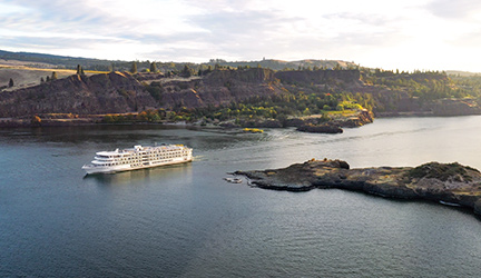 A small American Cruise Lines ship sails on a river, surrounded by scenic cliffs and lush greenery, under a partly cloudy sky.