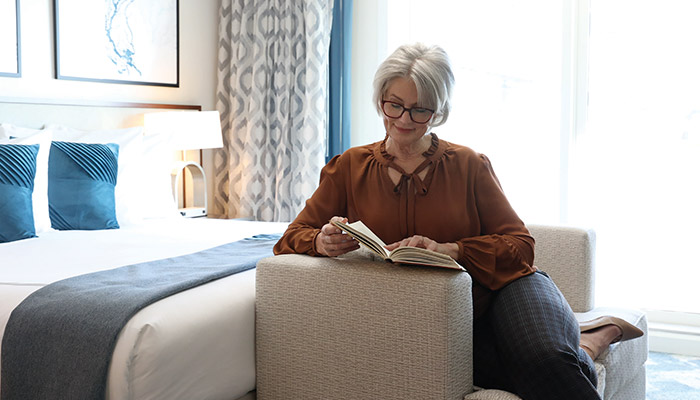 An older woman reading in a cozy American Cruise Lines ship stateroom with a spacious bed and blue accents, in front of a large window.