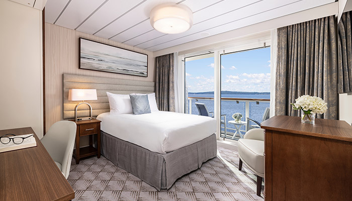 A solo American Cruise Lines ship stateroom with a single bed, desk, and chair, and a private balcony outside a large window.