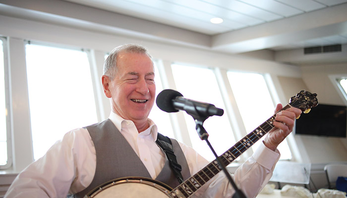 A male American Cruise Lines entertainer playing banjo and singing into a microphone, with large windows in the background.