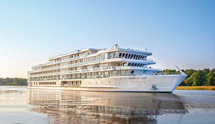 A small American Cruise Lines river ship sails on calm water, surrounded by lush greenery under a clear blue sky.