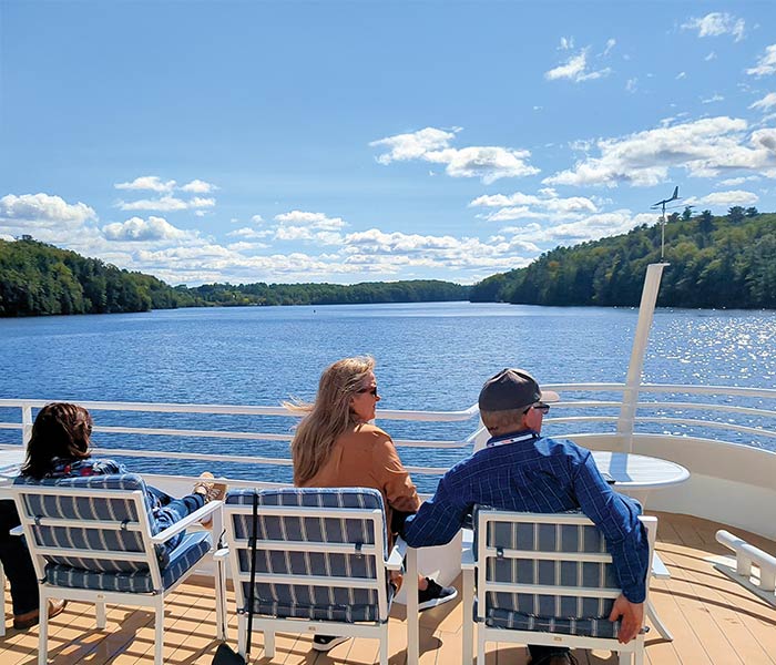 Three people seated on an American Cruise Lines ship deck, enjoying a scenic river view with lush green forests under a blue sky.