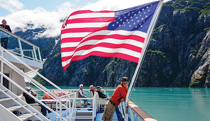 American Cruise Lines guests standing on deck beneath a U.S. flag while sailing through scenic American waterways.