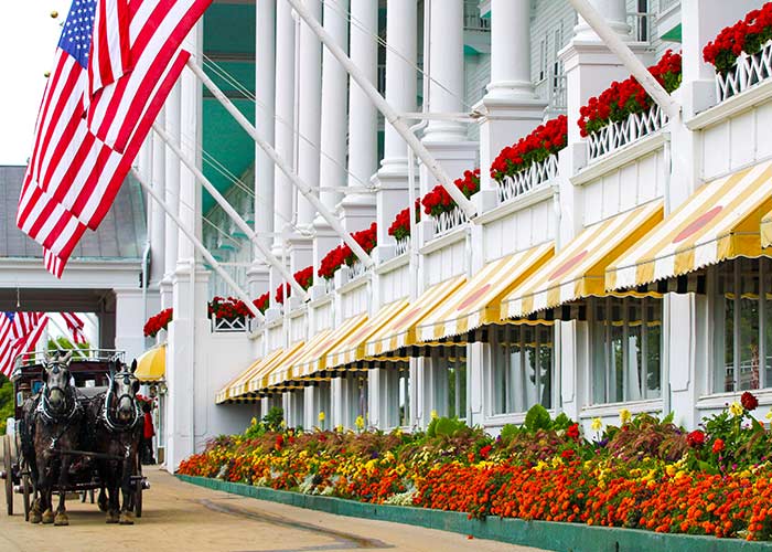 Horse-drawn carriage traveling past a white building with American flags and colorful flower beds on Mackinac Island.