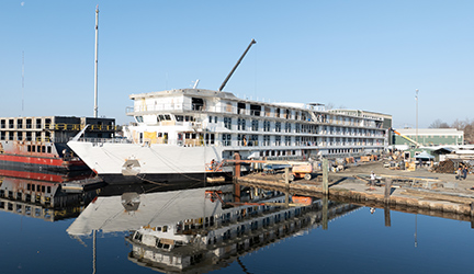 American Encore under construction at Chesapeake Shipbuilding, docked along the water with its reflection visible in the foreground.