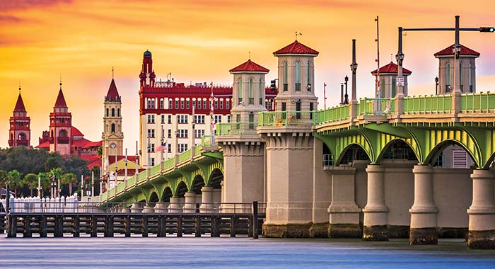 Green arched bridge spanning the water in St. Augustine under an orange sky at sunset, with historic buildings and towers in the background, seen near the city’s waterfront cruise port