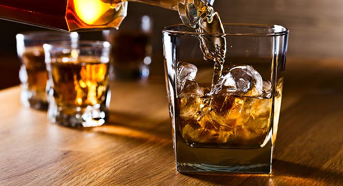 Whiskey being poured over ice into a glass on a wooden table with additional glasses in the background, at the St. Augustine Distillery.