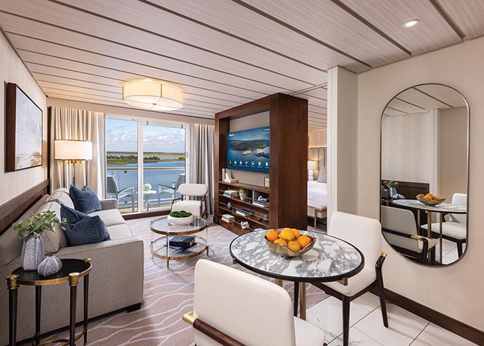 Spacious Grand Suite with sofa, chairs, round table, and floor-to-ceiling windows leading to a balcony with water views.