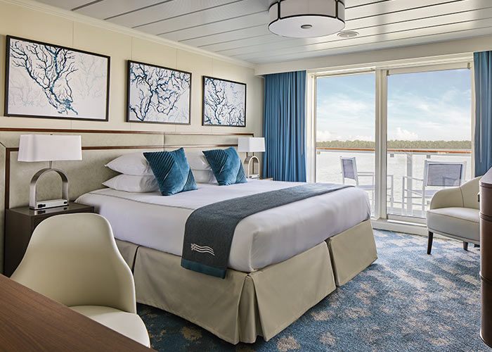 Stateroom with a large bed, side tables, seating area, and sliding glass doors opening to a balcony with water view.