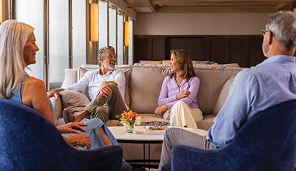 Four guests seated in an American Cruise Lines ship lounge, talking and smiling around a coffee table near large windows with natural light.